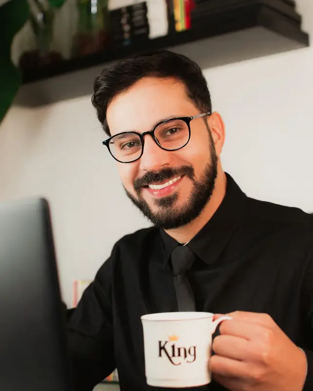 Thiago de óculos, sorrindo, com uma caneca na mão diante do notebook